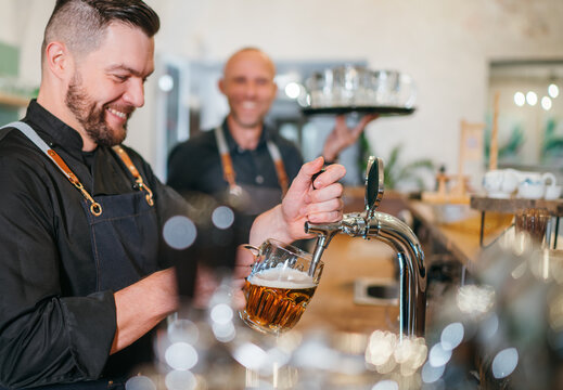 Stylish bearded barman dressed black uniform beer tapping at bar counter and waiter with tray chatting smiling to each other.Successful people team work friendship and good relationships concept image