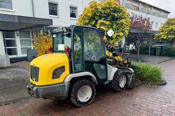 A small yellow backhoe loader standing on the sidewalk near residential buildings. Side view.