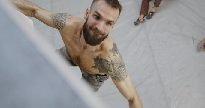 High angle view of a fit young man with tattoos climbing up an artificial wall in a bouldering gym with people watching from below