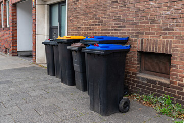 Plastic trash cans with colorful lids stand on the sidewalk near a brick wall of a building.
