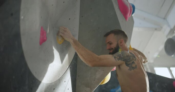 Fit young man with tattoos using handholds to climb up an overhang on rock wall in a bouldering gym