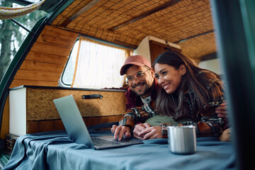 Happy couple of campers surfing the net on laptop on their van in woods.
