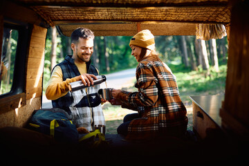 Happy couple drinking tea while camping with their van in woods.