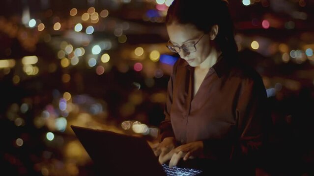 Young woman works on laptop in dark on background bokeh of night city lights. business woman working late