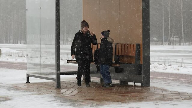 Mother and son warm up at public transport stop on frosty winter day. Family runs and jumps while waiting for bus in snowfall. Discomfort of living at sub-zero temperatures.