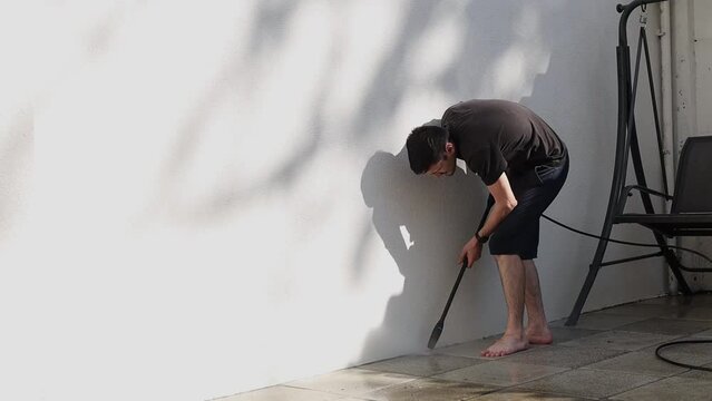One young Caucasian brunette man in dark clothes stands bending over and washes the grout of dirty garden tiles near a white wall with a Karcher, on a summer evening in the small backyard of his house
