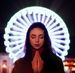 young woman with closed eyes puts her hands together in front of a ferris wheel