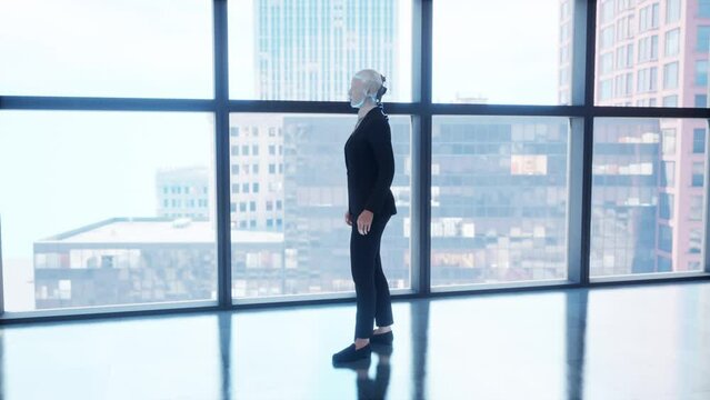 A robot humanoid staying in the empty office and looking out the window at the big city. future technology concept