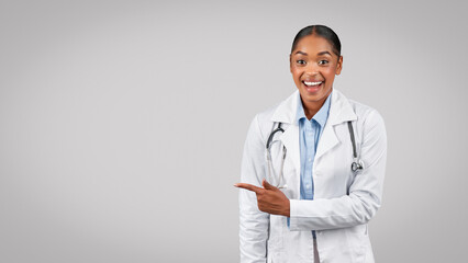 Shocked glad millennial black lady doctor point finger to free space, isolated on gray background