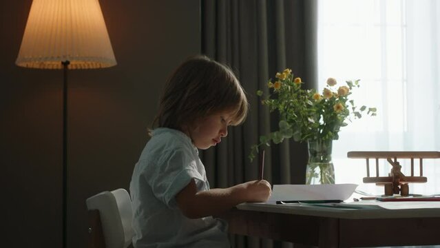 Diligent boy draws against background of window at table. Cinematic. Kaleidoscope of emotions, essence of childhood. Innermost thoughts and inspirations. Happy childhood concept, calm measured life