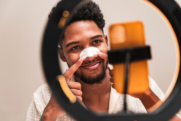 A cheerful young latin man applies a pore-cleansing nose strip while looking in the mirror, highlighting a personal grooming moment.