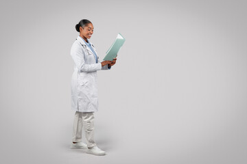 Cheerful black millennial lady doctor in white coat, with clipboard, look at free space