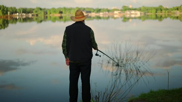Grandfather fishing on local lake at the village. Alone grandpa catching fishes outdoors. Rear view of senior man holding fish rod in nature environment. Retired fisherman calmness