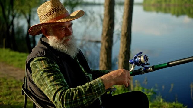 Concept of favorite hobby. Relaxing pensioner sitting in chair when fishing near the river. Satisfied grandfather handle spinning fish rod outdoors. Retired man angler