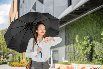 A young Asian businesswoman, shielded by an umbrella, walks to the office under hot sunlight,...