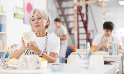 Talented senior female artisan in apron working on embellishing ceramic mug at table in modern pottery studio, applying design with paints.. - obrazy, fototapety, plakaty