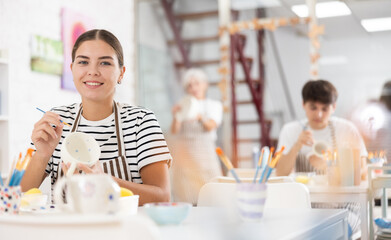Positive young girl attending pottery class, enthusiastically painting on ceramic mug, creating...
