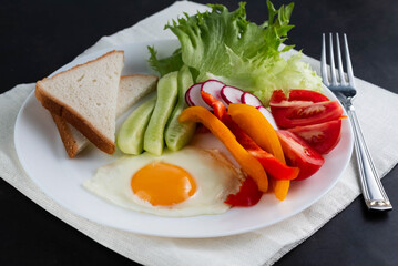 On a white plate there is an egg, slices of pepper, tomatoes, cucumbers, radishes and a leaf of green salad with a fork on the side on a dark table.