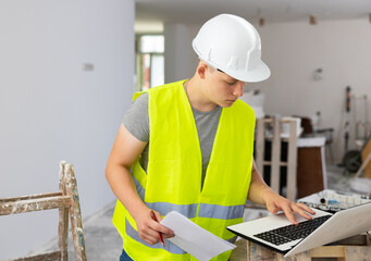 Young man using laptop and verifying papers on part time job in construction site.