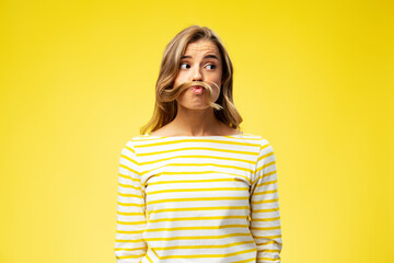 Portrait of happy young woman playing with hair, showing grimace looking away isolated on yellow background. Concept of positive lifestyle