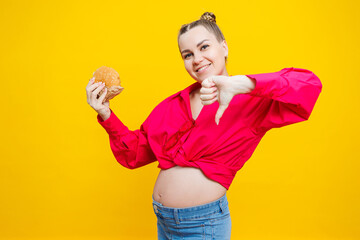 A cute pregnant woman in a pink shirt is eating a fast food burger. A pregnant woman on a yellow background with an appetizing burger in her hands. Harmful food for pregnant women.