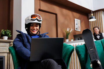 Asian female tourist wearing winter clothing enjoying her time on sofa with digital device in hotel lobby. Excited woman with skiing equipment browsing on laptop for slopes at ski mountain resort. - obrazy, fototapety, plakaty