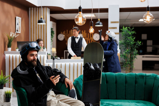 Traveler dressed in winter clothing waits in hotel lobby, browsing on smartphone. Male tourist checks ski resort reservation on mobile phone while seated on couch. Receptionist assists another guest.