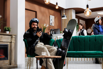 A man seated on sofa browses on his digital phone with snowboard nearby. Caucasian visitor dressed in snow clothing surfing the net using his smartphone. Winter vacation anticipation captured.