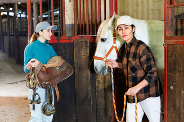 Women ranchers preparing white horse for ride. Asian woman leading horse, European woman carrying...