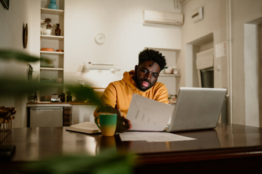 A focused interracial casual financial adviser is looking at the statistics and paperwork while sitting at home with a laptop and working remotely.