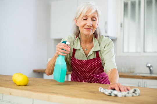 Smiling senior woman in apron washing top of kitchen table during cleanup in apartment.