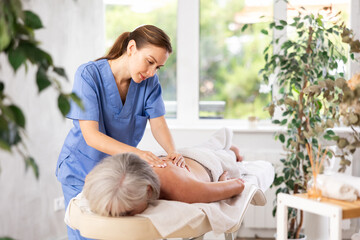 Young female masseuse doing back massage to elderly woman client in massage room