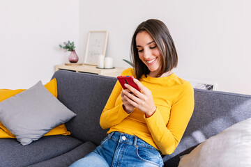 Happy and smiling beautiful young woman using smart phone device sitting on sofa in the living...