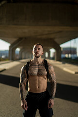 Chico joven tatuado y musculoso posando y haciendo deporte en la calle en un tunel con un puente de fondo