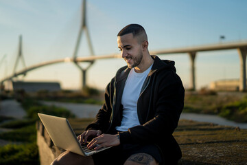 Chico joven emprendedor trabajando en su laptop al atardecer con un puente de fondo 