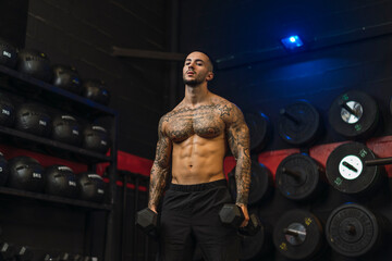 Chico joven tatuado y musculoso posando con iluminación de estudio en gimnasio de crossfit