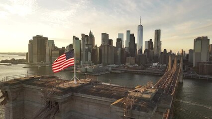 Aerial shot of The American Flag flying high above New York City's Brooklyn Bridge at sunset.