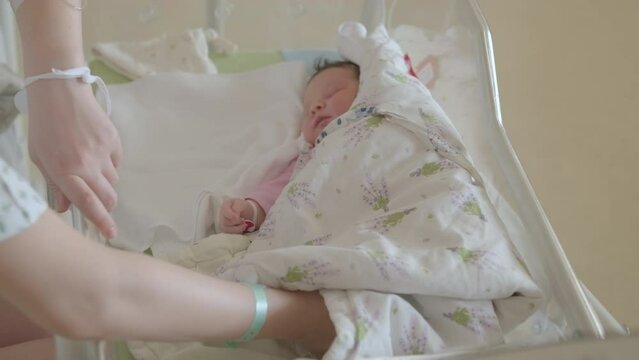 Mother stands near crib with newborn daughter in hospital with hands inside. Child shows slow movements while wrapped in blanket lying peacefully
