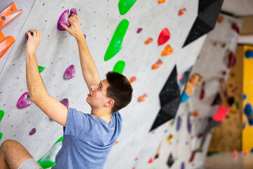 European young man grabbing ledges of artificial climbing wall in bouldering centre. - obrazy, fototapety, plakaty