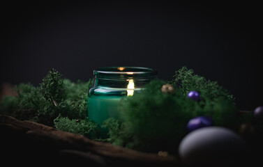 A burning candle in a glass jar in the bark of a tree with moss and precious ritual stones on a...