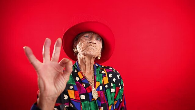 Funny portrait of mature elderly woman, 80s, having giving OKAY gesture, wearing red hat isolated on red background.