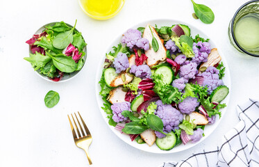 Healthy fresh salad from purple cauliflower, grilled chicken breast, cucumbers, red onion, radicchio, spinach with pumpkin seeds, white table background, top view