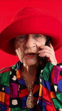 Vertical video of funny closeup portrait of toothless old elderly woman wearing red hat isolated on red background talking on cell phone.