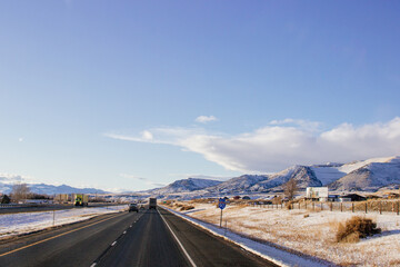 A beautiful landscape with mountains covered in snow, a highway with cars driving along it, and a blue sky with fluffy clouds on a sunny winter day. Butte, Montana, USA