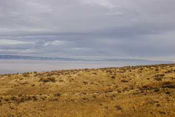 An oblique panorama with dry grass in the foreground, a landscape with roads, fields, meadows, mountains that are hidden in clouds and fog on the horizon. Picturesque landscape with beautiful sky