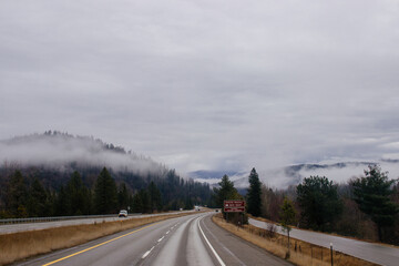 Asphalt road in the middle of high mountains, covered with fog and clouds, at dusk. American winter landscape of a mountainous area covered with fir forest
