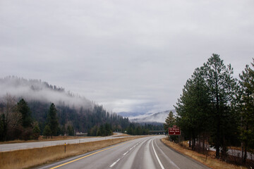 Obraz premium Asphalt road in the middle of high mountains, covered with fog and clouds, at dusk. American winter landscape of a mountainous area covered with fir forest