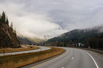 Asphalt road in the middle of high mountains, covered with fog and clouds on autumn day. American winter landscape of a mountainous area covered with fir forest. Fall season on the highway with cars