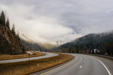 Asphalt road in the middle of high mountains, covered with fog and clouds on autumn day. American winter landscape of a mountainous area covered with fir forest. Fall season on the highway with cars