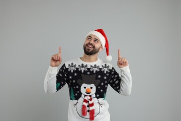Happy young man in Christmas sweater and Santa hat pointing at something on grey background
